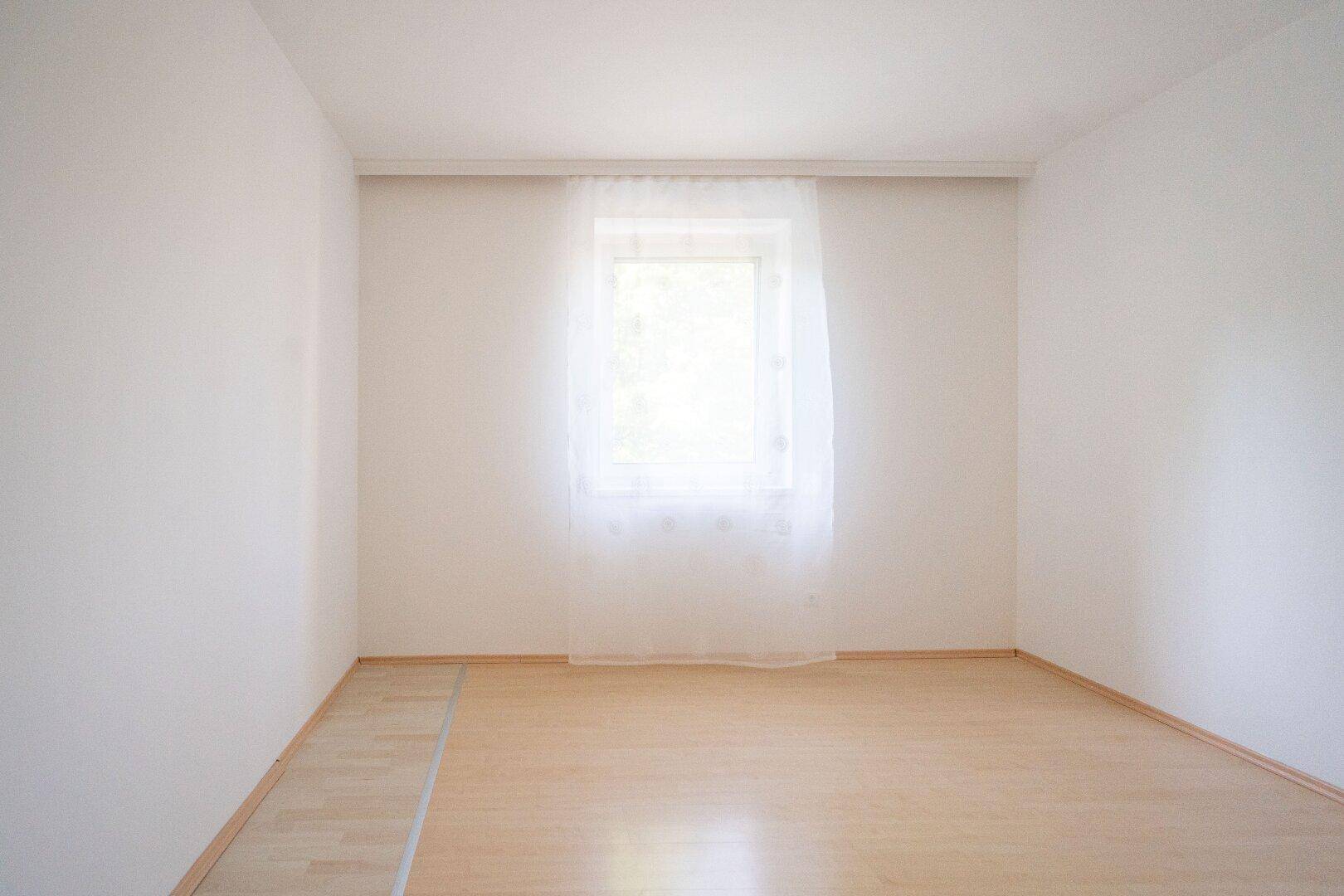 Leeres Zimmer mit hellem Laminatboden und einem Fenster, das viel Tageslicht hereinlässt.