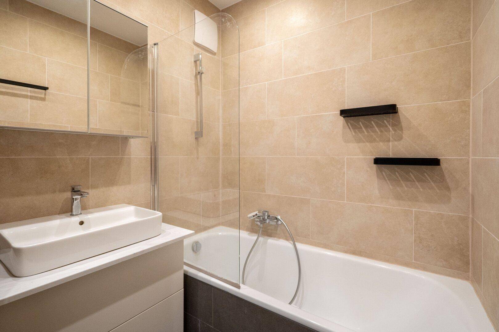 Modernes Badezimmer mit beigefarbenen Fliesen, Badewanne mit Duschwand und Waschtisch.