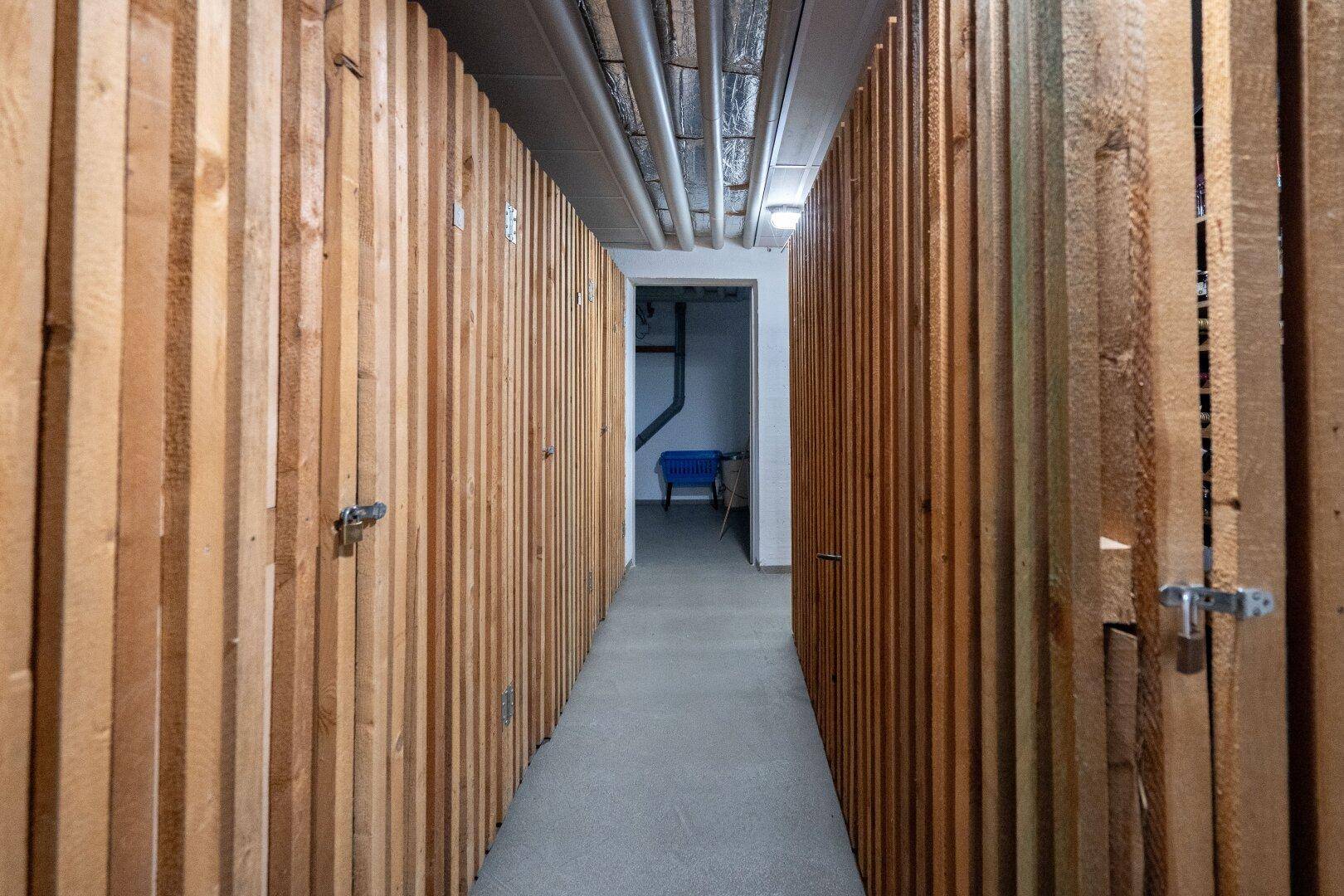 Kellergang mit individuellen Holzverschlägen für zusätzlichen Stauraum und sichtbaren Rohren.