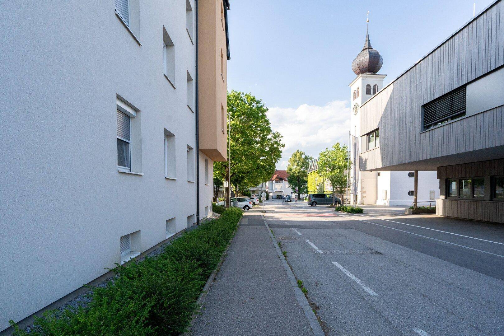 Straßenansicht mit Wohngebäuden, Bürgersteig und einem Kirchturm in der Ferne unter blauem Himmel.