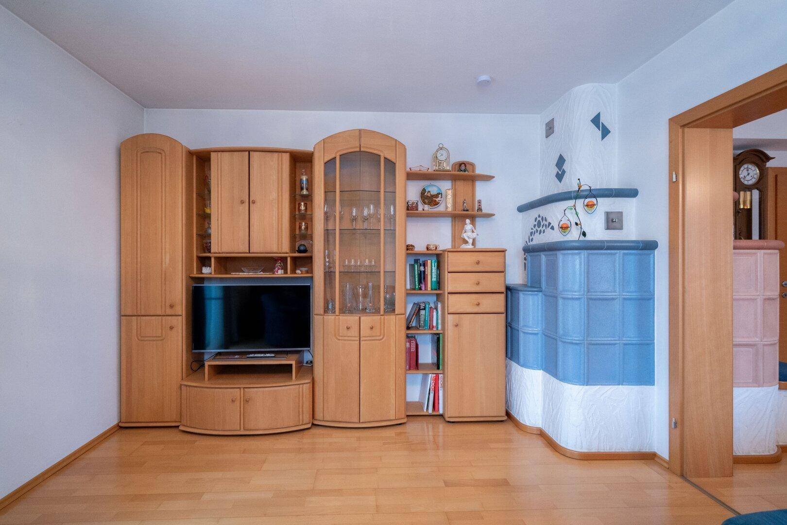 Wohnzimmer mit hellem Holzboden, großer Schrankwand und blauem Kachelofen.