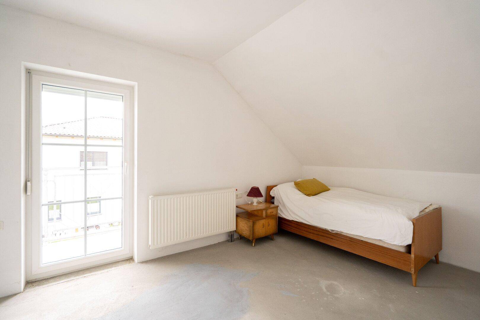 Dachgeschoss-Schlafzimmer im Rohbau mit Bett, Fenster und Heizkörper.