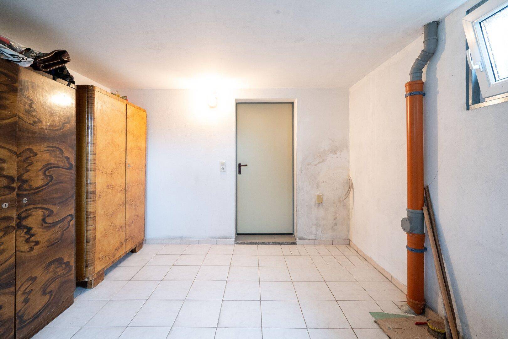 Geräumiger Kellerraum mit Fliesenboden, Holzschrank und sichtbaren Leitungen.