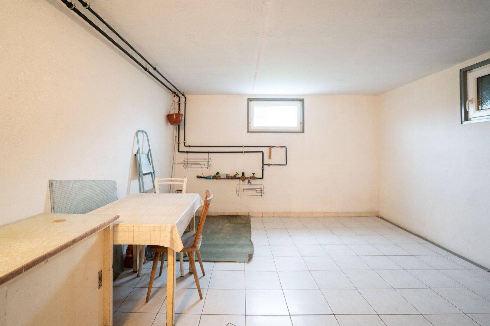 Heller Kellerraum mit Fliesenboden, Tisch und Stühlen sowie sichtbaren Rohrleitungen.