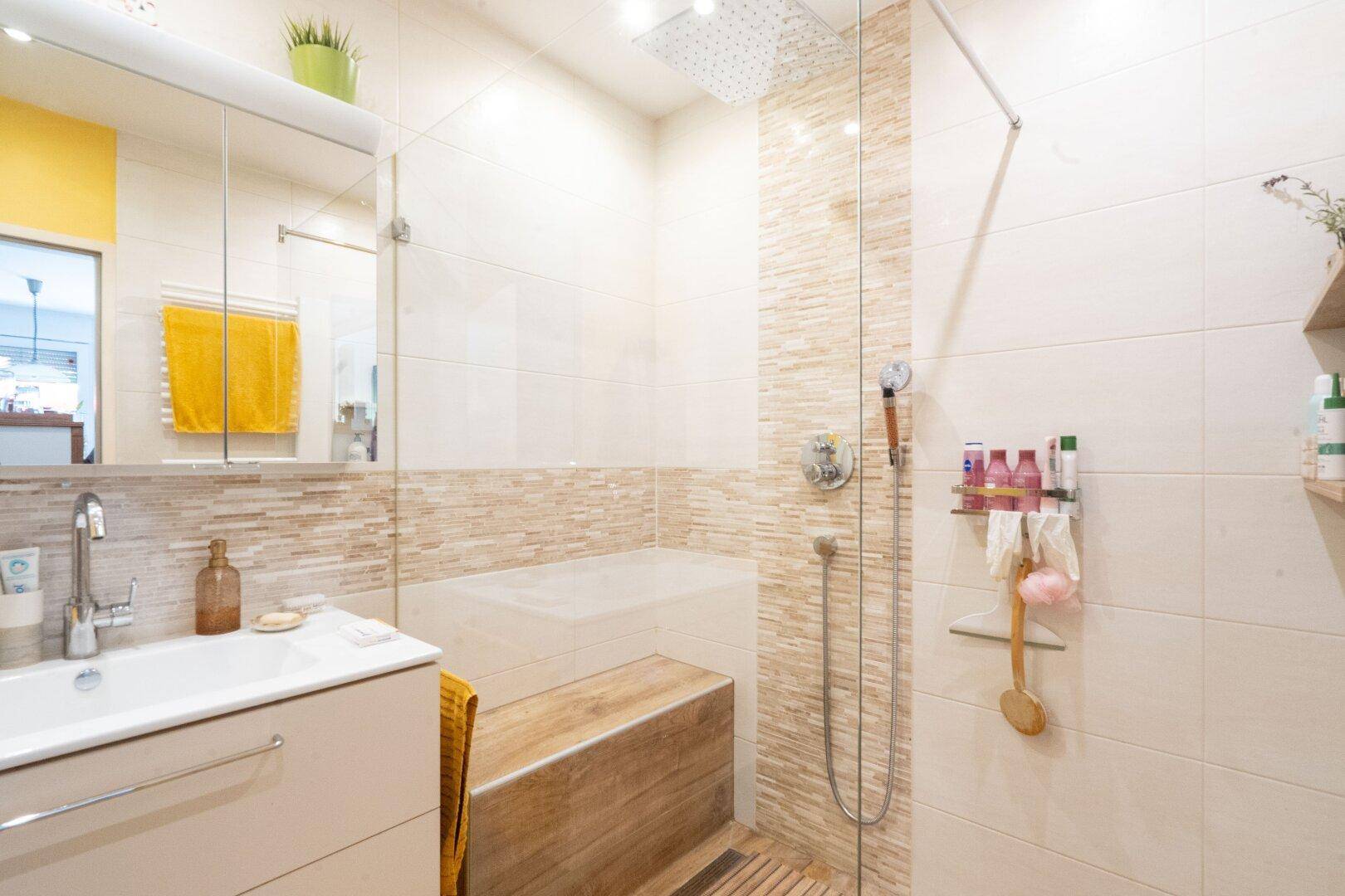 Modernes Badezimmer mit begehbarer Dusche, Holzbank und hellen Fliesen.