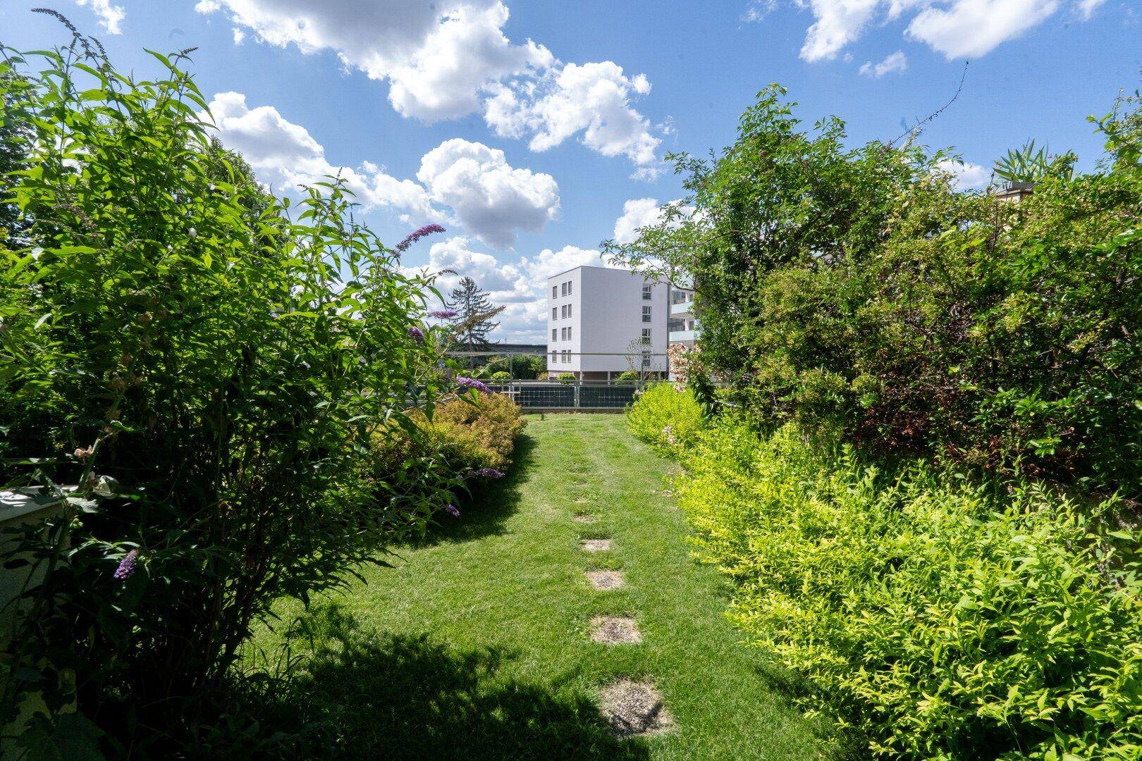 Grüner Gartenweg mit Trittsteinen, umgeben von Büschen und Blick auf ein Gebäude.