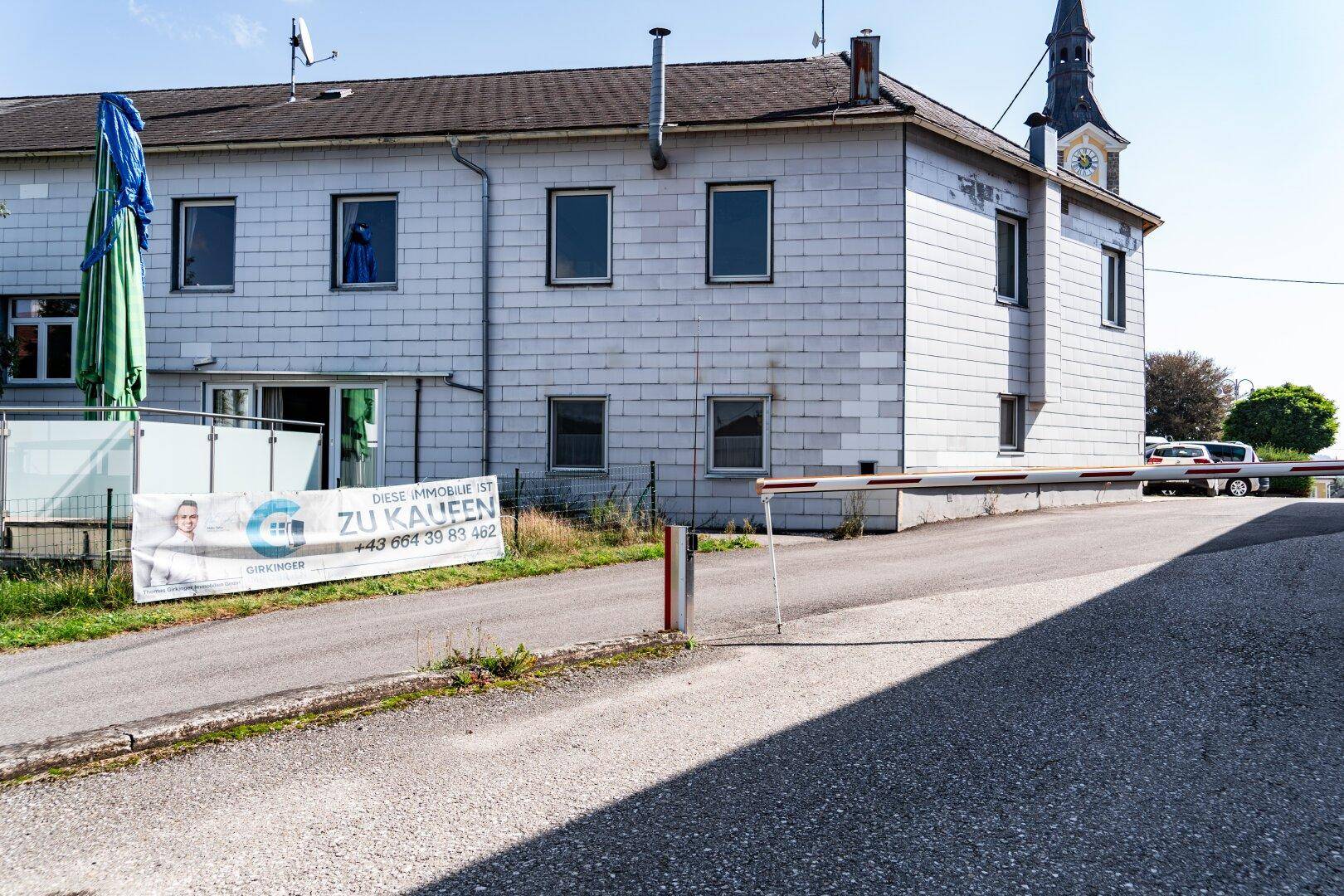 Graues Gebäude mit weißer Fassade und einem "ZU KAUFEN" Banner, umgeben von einer Schranke und Grünflächen.