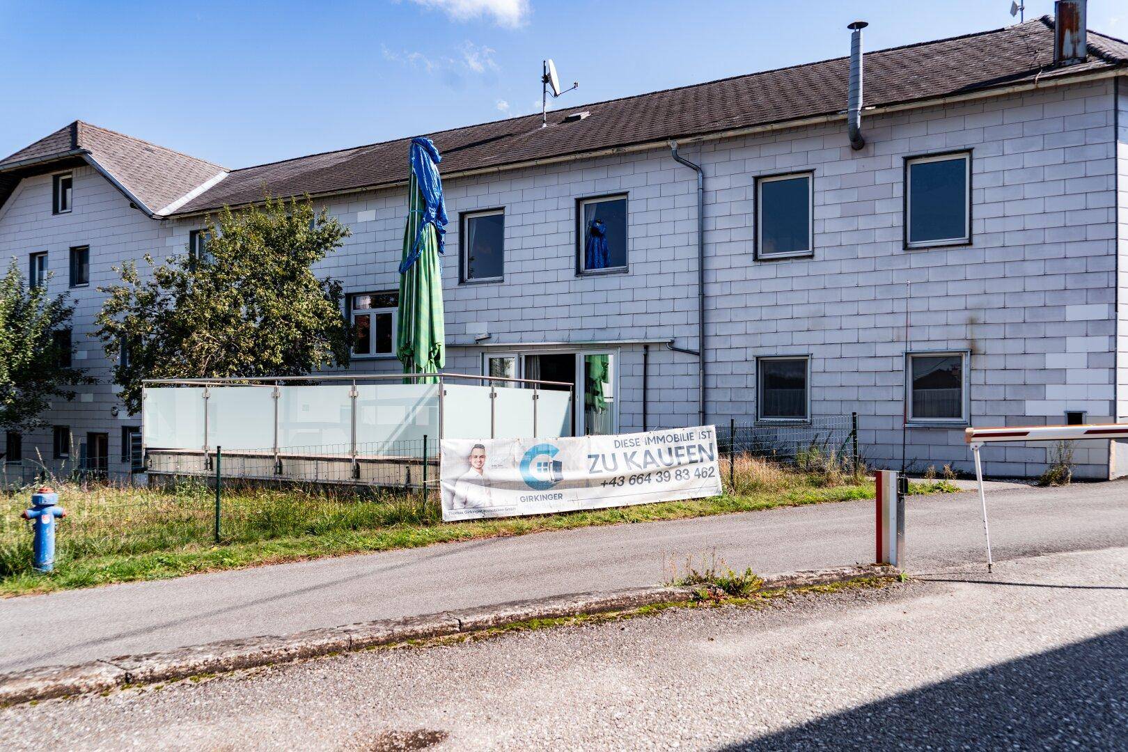 Ansicht eines grauen Gebäudes mit einem großen grünen Sonnenschirm und einem "ZU KAUFEN" Banner.