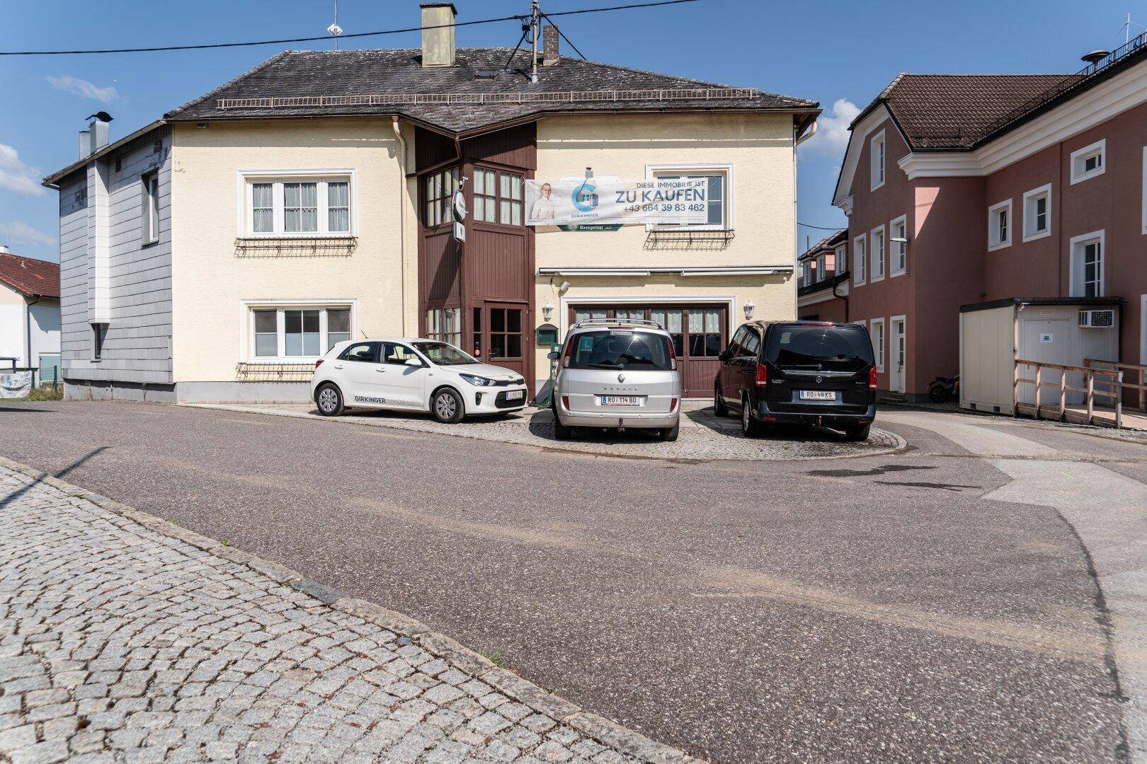 Gelbes Gebäude mit "ZU KAUFEN" Banner an einer Straße mit geparkten Autos unter blauem Himmel.