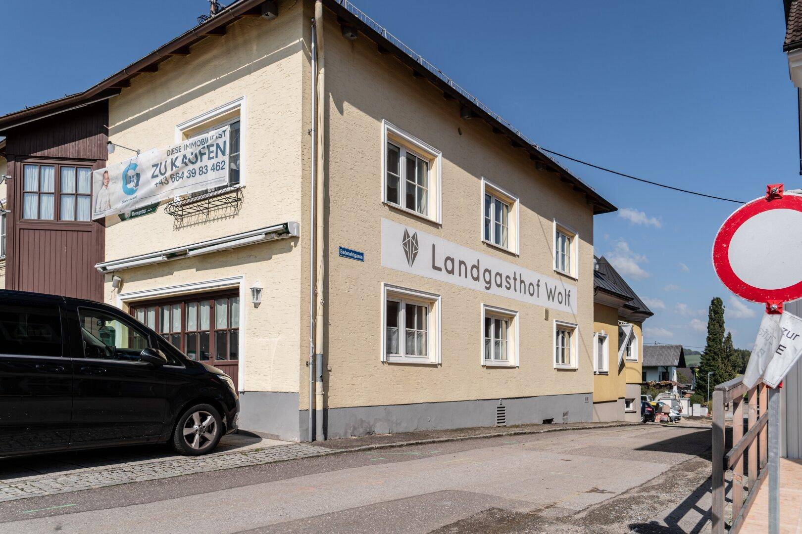 Seitenansicht eines gelben Gebäudes mit "Landgasthof Wolf" Schriftzug und einem "ZU KAUFEN" Banner.