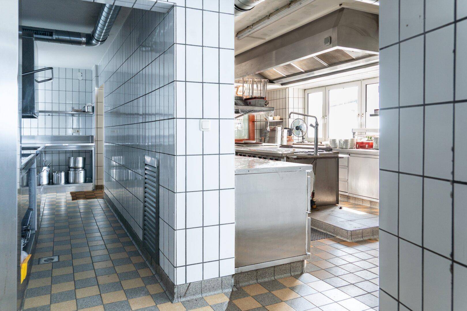 Blick in eine professionelle Küche mit Edelstahlgeräten, gefliesten Wänden und einem Fenster für Tageslicht.