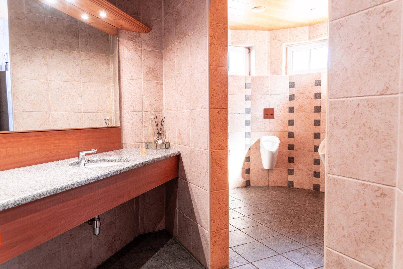 Öffentliche Toilette mit mehreren Urinalen, langem Waschtisch mit Granitplatte und gefliesten Wänden.