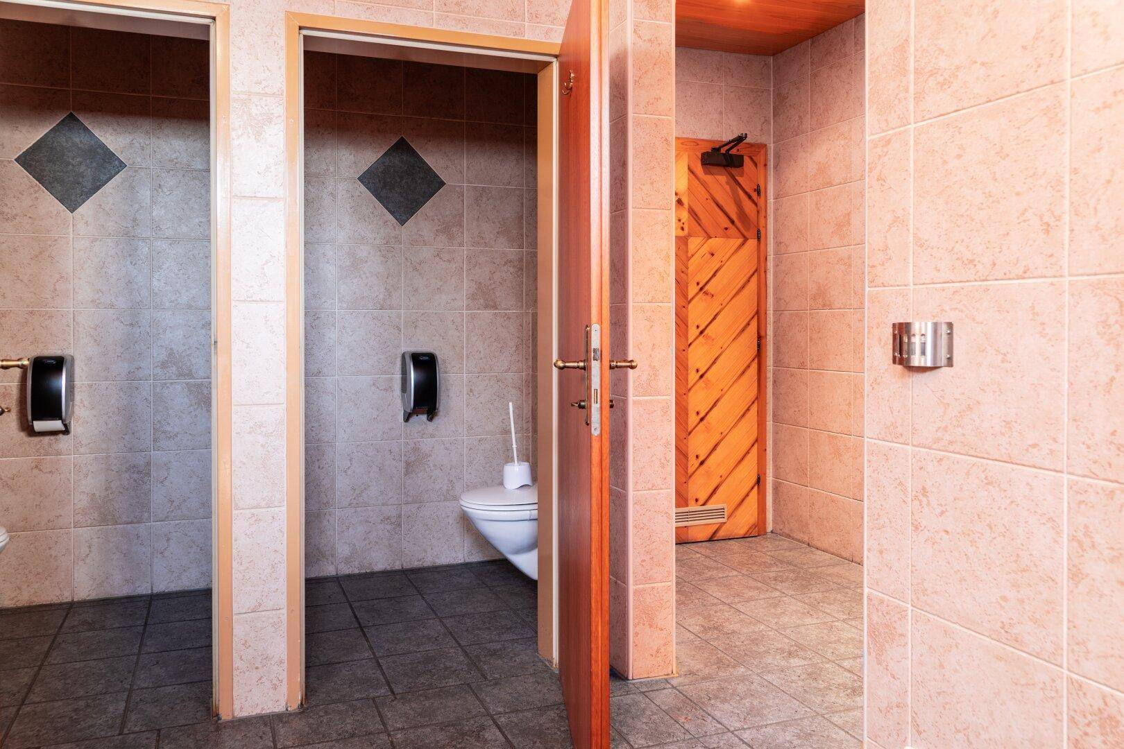 Öffentliche Toilette mit mehreren Toilettenkabinen, gefliesten Wänden und einer Holztür.