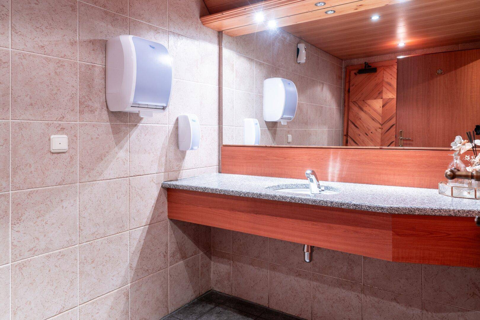 Öffentliche Toilette mit langem Waschtisch, Granitplatte, Waschbecken, Spiegel und gefliesten Wänden.