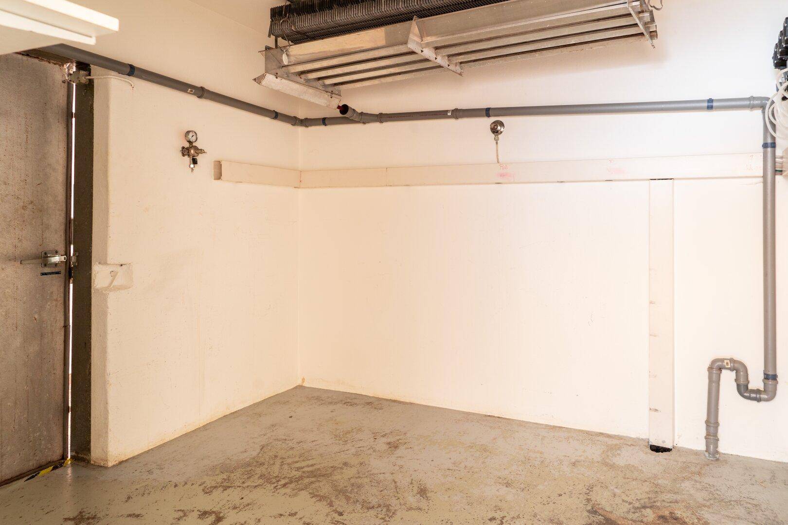 Leerer Raum mit weißen Wänden, Betonboden und sichtbaren Rohren an Decke und Wand.