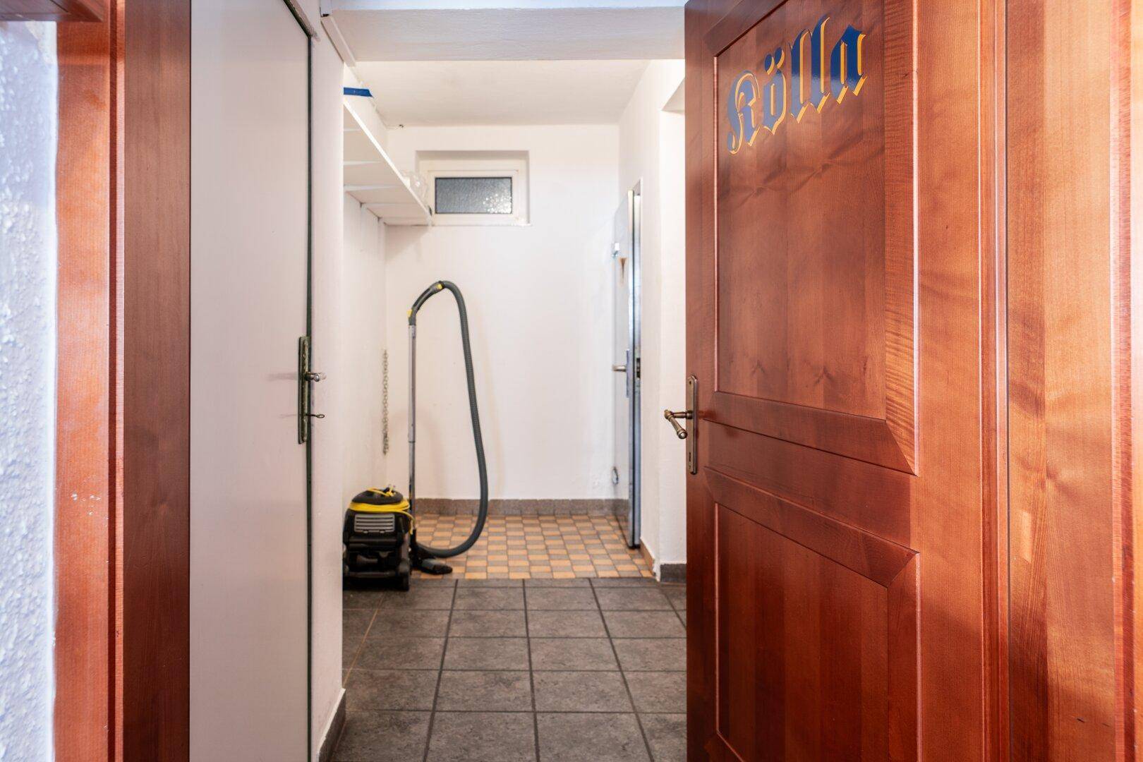 Flur oder Hauswirtschaftsbereich mit Staubsauger, Fliesenboden und Holztüren, kleines Fenster.