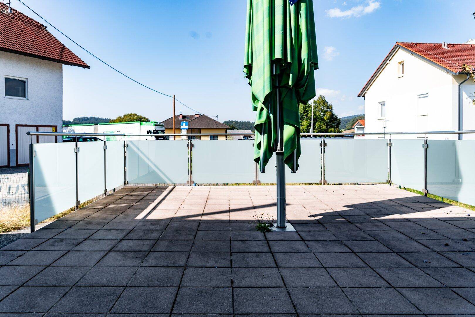 Große gepflasterte Terrasse mit Glasgeländer und einem zusammengeklappten Sonnenschirm unter blauem Himmel.