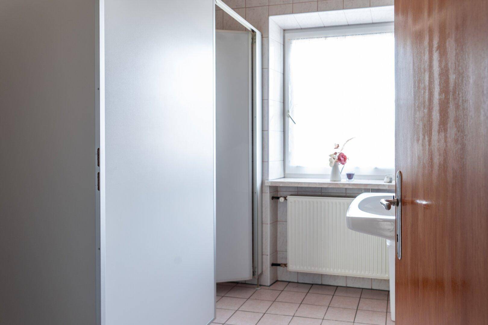 Badezimmer mit Waschbecken, Fenster, Heizkörper und Duschkabine, hell und funktional gestaltet.