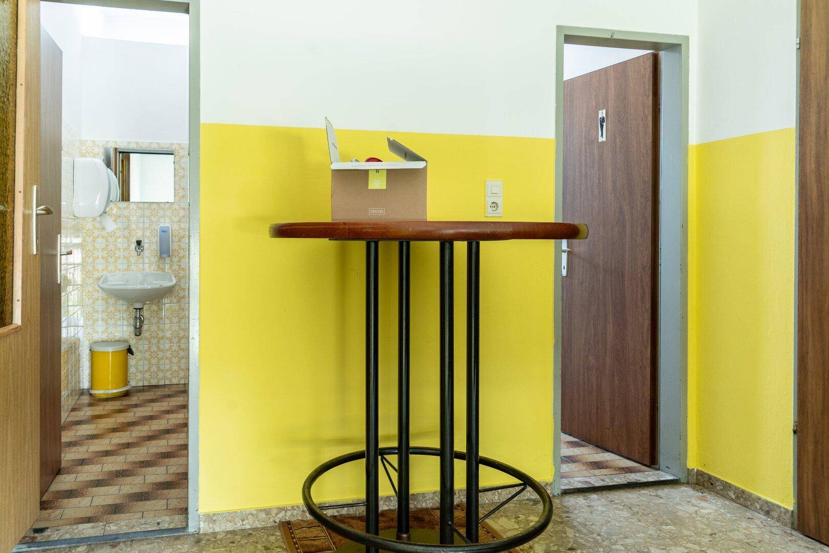 Flur mit Stehtisch, der zu zwei separaten Räumen führt, wahrscheinlich Toiletten, mit gelb gestrichenen Wänden.