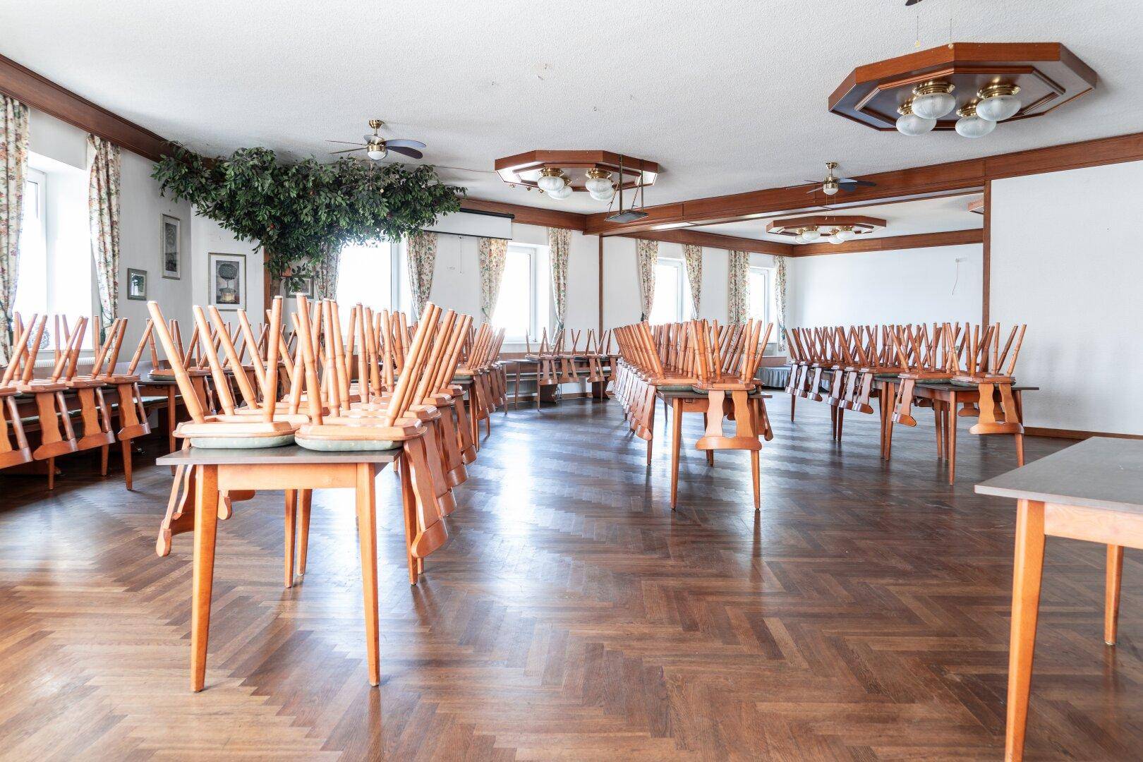 Großer Speisesaal oder Veranstaltungsraum mit gestapelten Holztischen und Stühlen, Parkettboden und Deckenventilatoren.