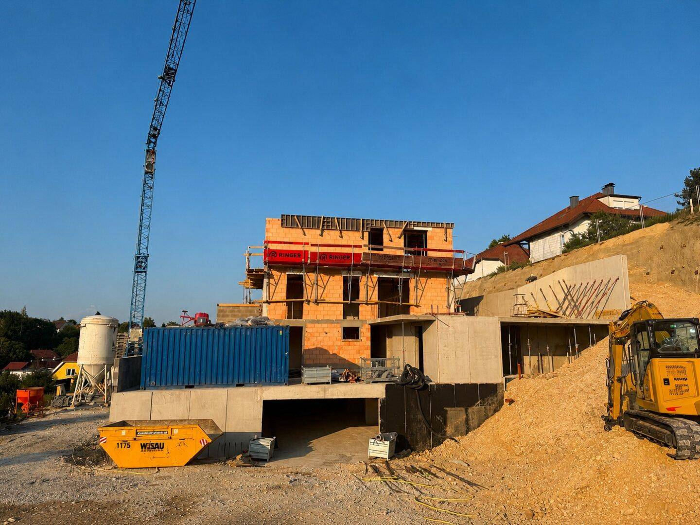 Baustelle mit im Bau befindlichem Gebäude, Bagger und Containern auf einem Hanggrundstück.