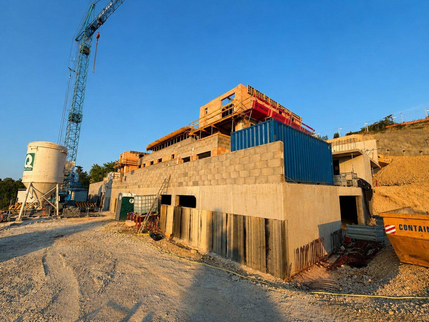 Baustelle eines Neubaus mit Kran, Containern und Erdarbeiten an einem Hanggrundstück.