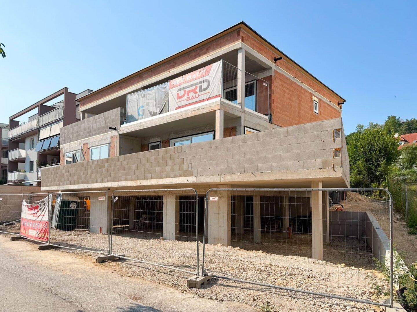 Vorderansicht des Neubaus im Rohbau mit Bauzäunen und Werbebannern.