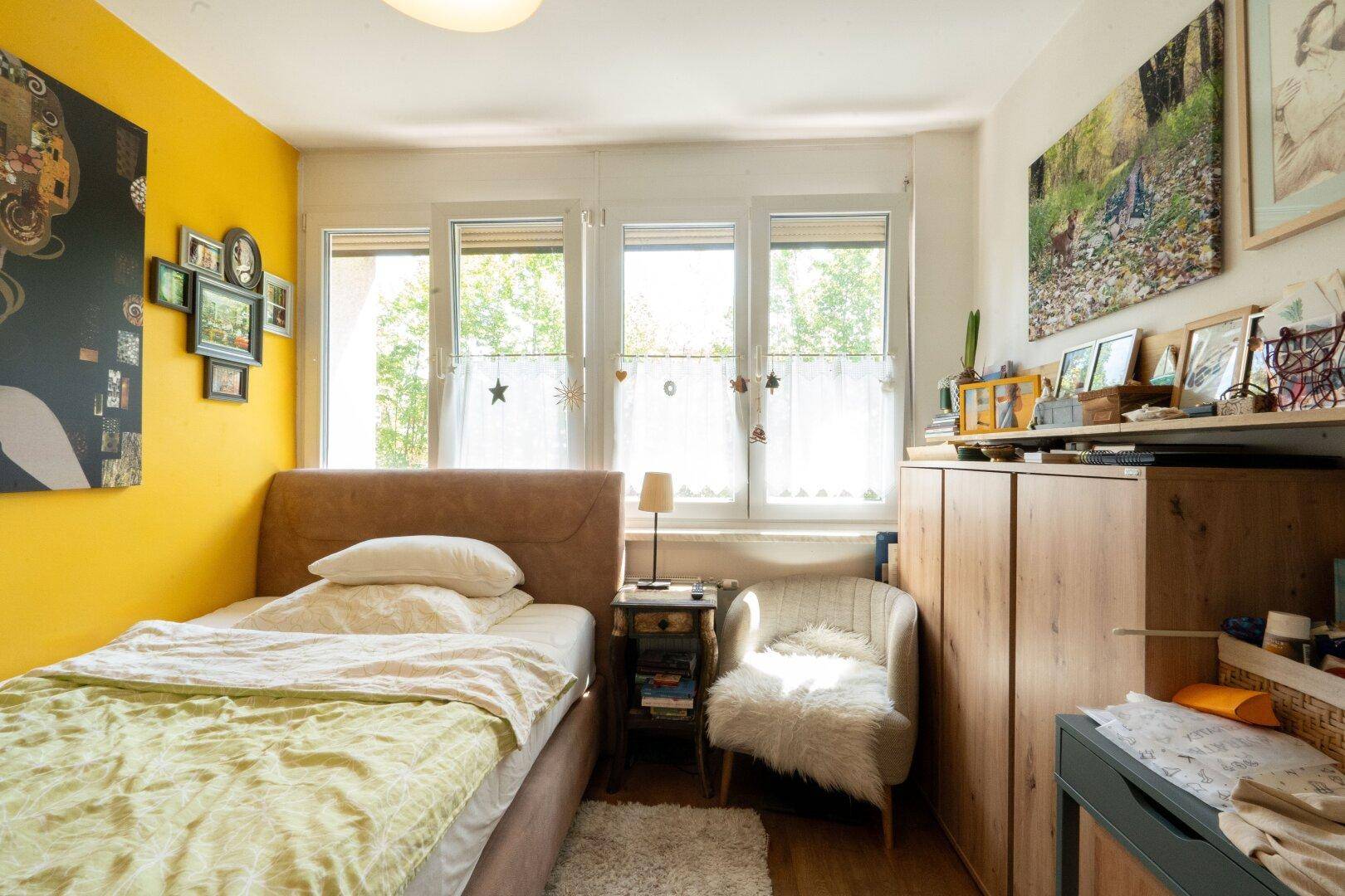 Gemütliches Schlafzimmer mit Einzelbett, gelber Akzentwand und großem Fenster für viel Tageslicht.