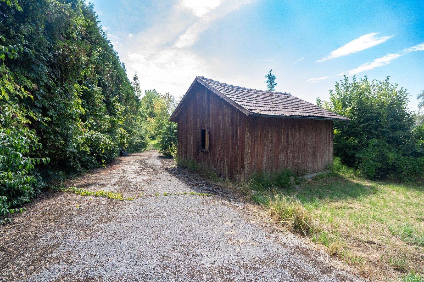 Rustikaler Holzschuppen mit Ziegeldach neben einem Schotterweg, umgeben von Bäumen und Gras.