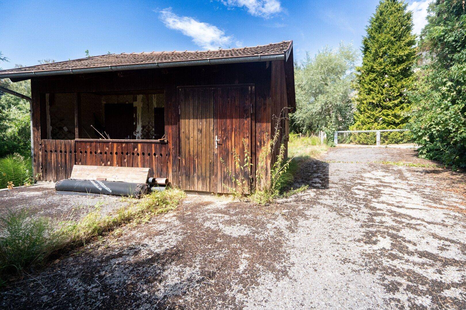 Rustikaler Holzschuppen mit offener Seite und geschlossener Tür, daneben eine Schotterfläche.