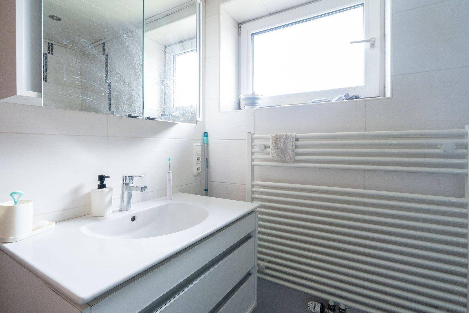 Modernes Badezimmer mit Waschbecken, Spiegelschrank, Fenster und Handtuchheizkörper.