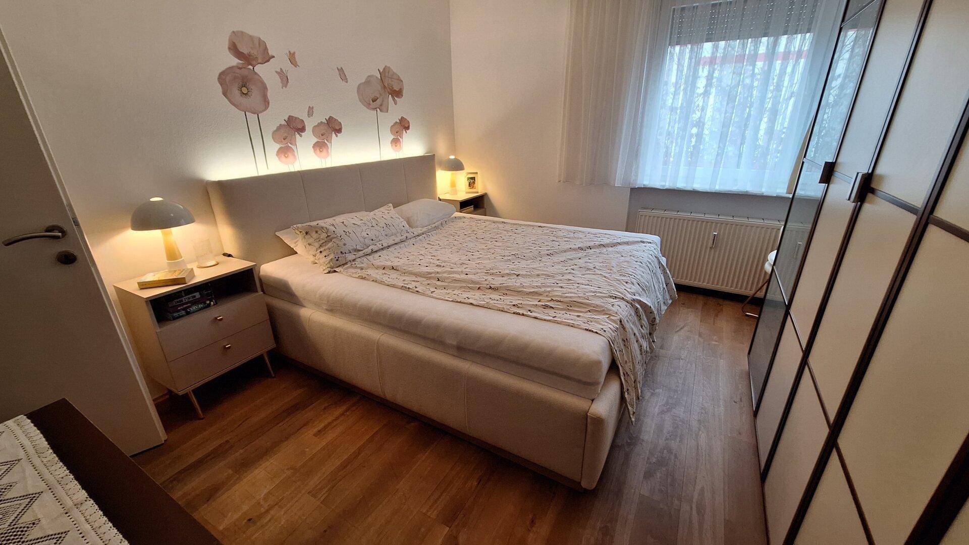 Ruhiges Schlafzimmer mit Doppelbett, Holzfußboden und dezenter floraler Wanddekoration für eine entspannte Atmosphäre.