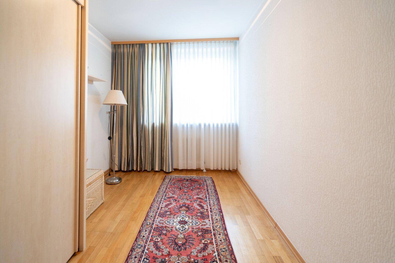 Schmaler Raum mit Holzfußboden, Teppichläufer und einem Fenster mit Vorhängen.