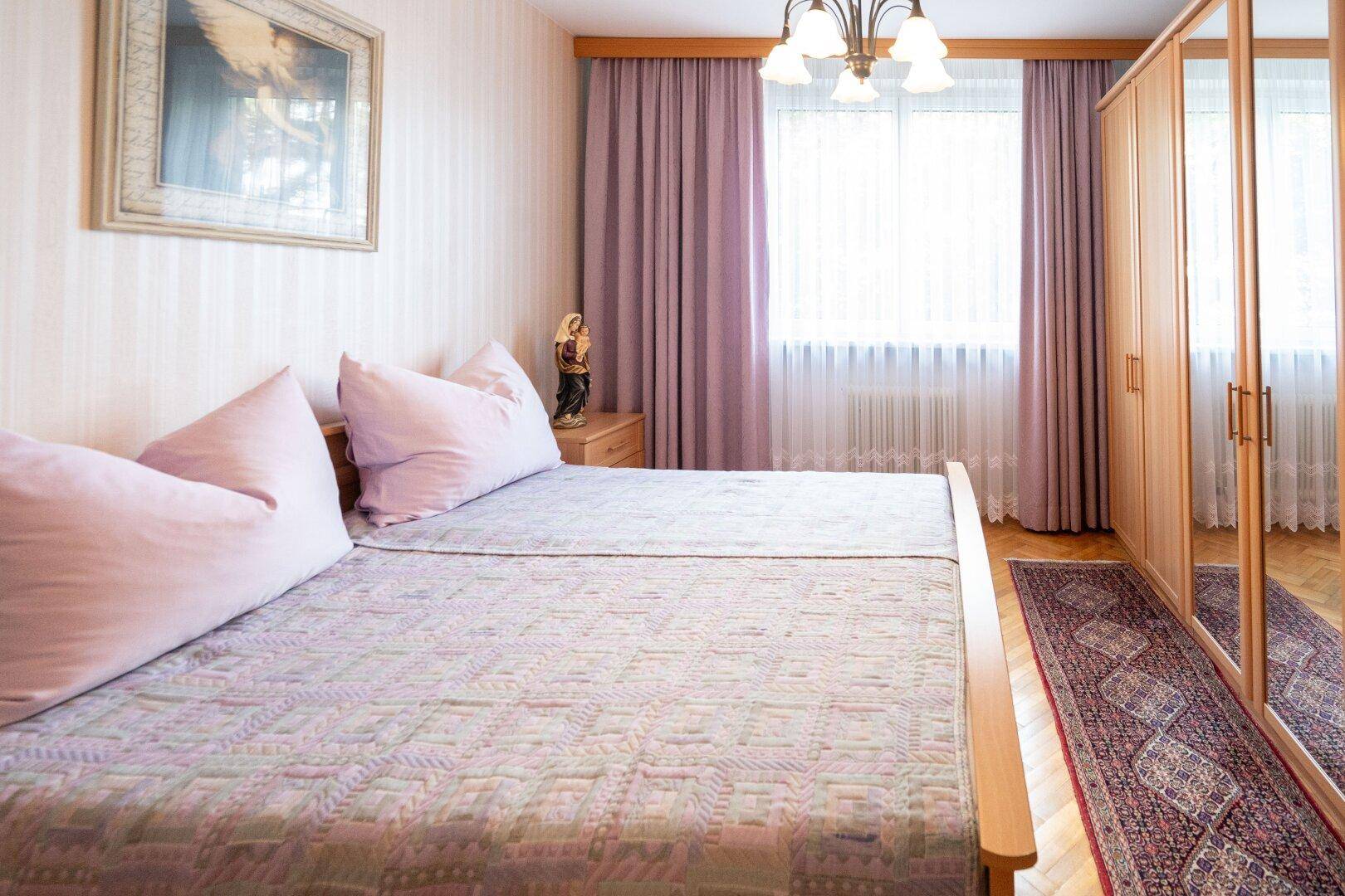 Schlafzimmer mit Doppelbett, Spiegelschrank und hellen Vorhängen für viel Tageslicht.