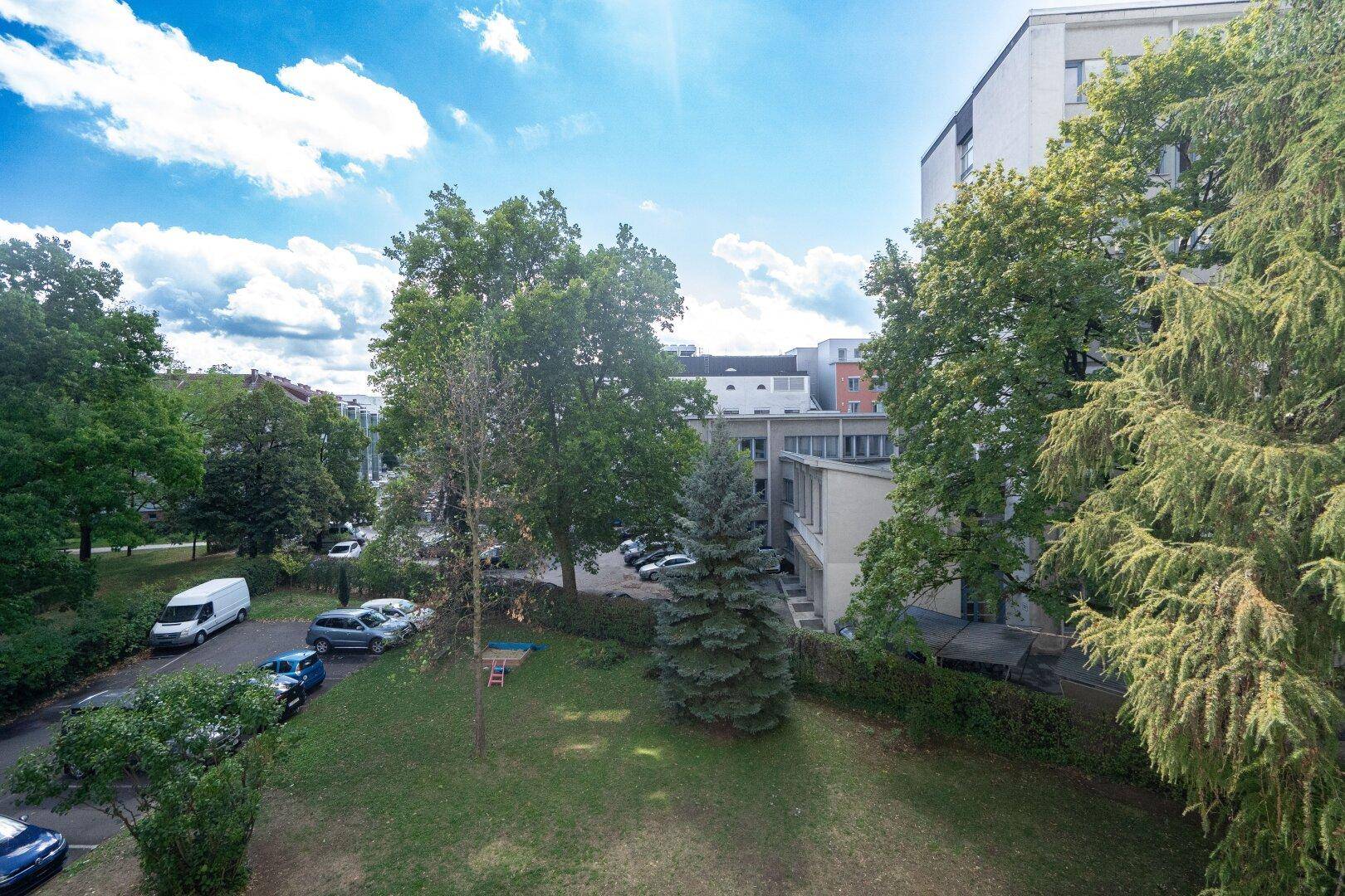 Außenansicht mit Bäumen, Parkplätzen und umliegenden Gebäuden unter blauem Himmel.