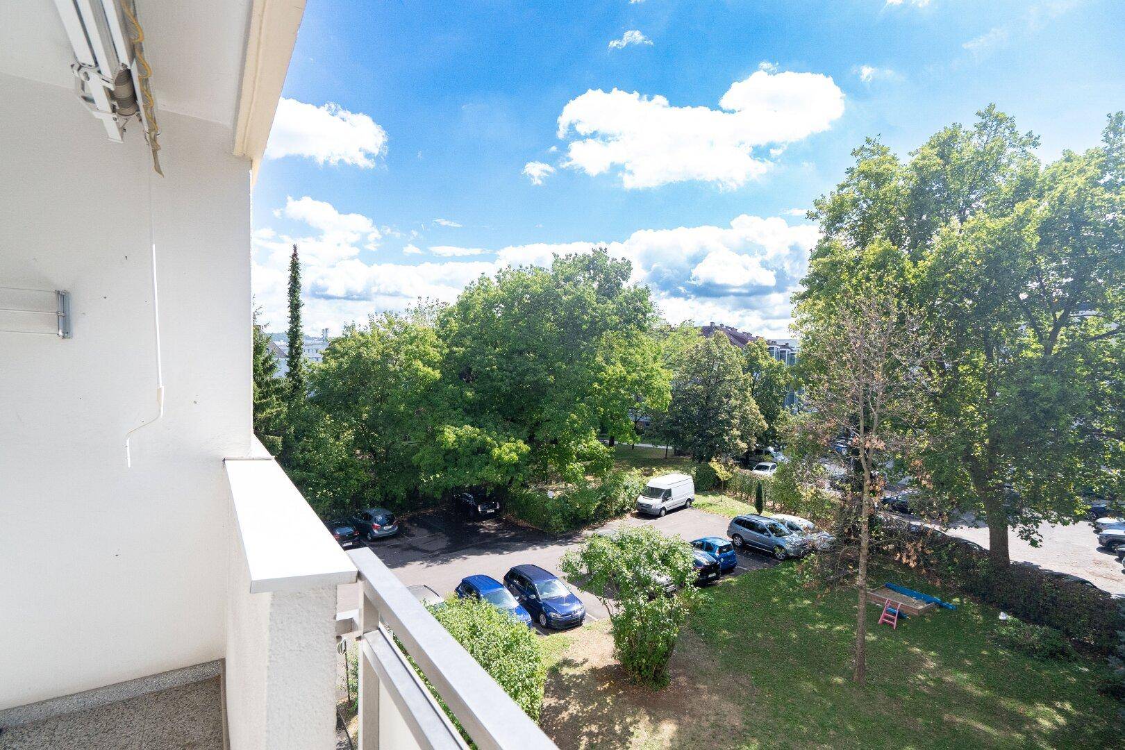 Weitblick vom Balkon auf grüne Bäume und Parkplätze unter blauem Himmel.