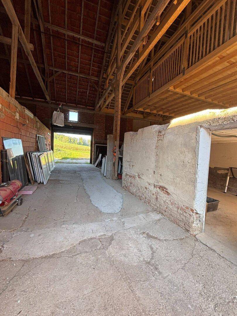 Innenansicht eines Lagerraums mit Holzbalken, Betonboden und einer Rampe, die zum Ausgang mit Feldblick führt.