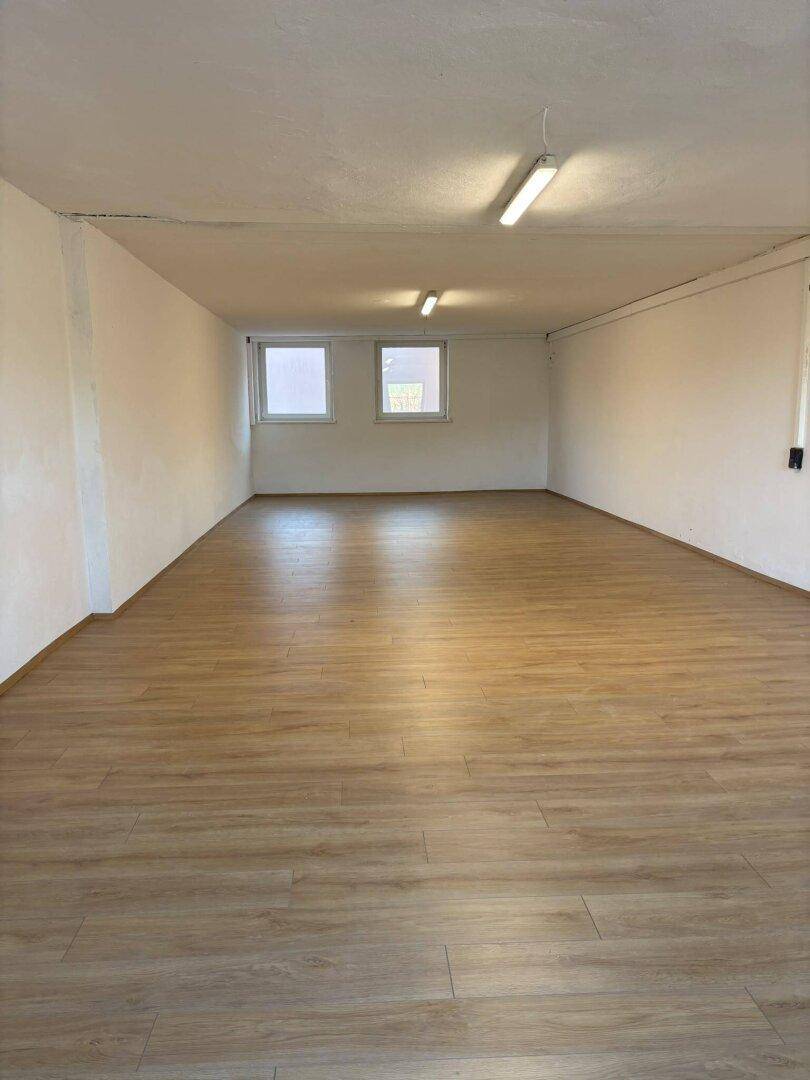Leerer Raum mit hellem Holzboden und weißen Wänden, beleuchtet durch zwei Fenster und Deckenleuchten.