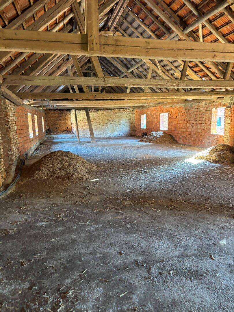 Großer, leerer Innenraum eines alten Gebäudes mit Holzbalken, Ziegelwänden und Heuhaufen auf dem Boden.