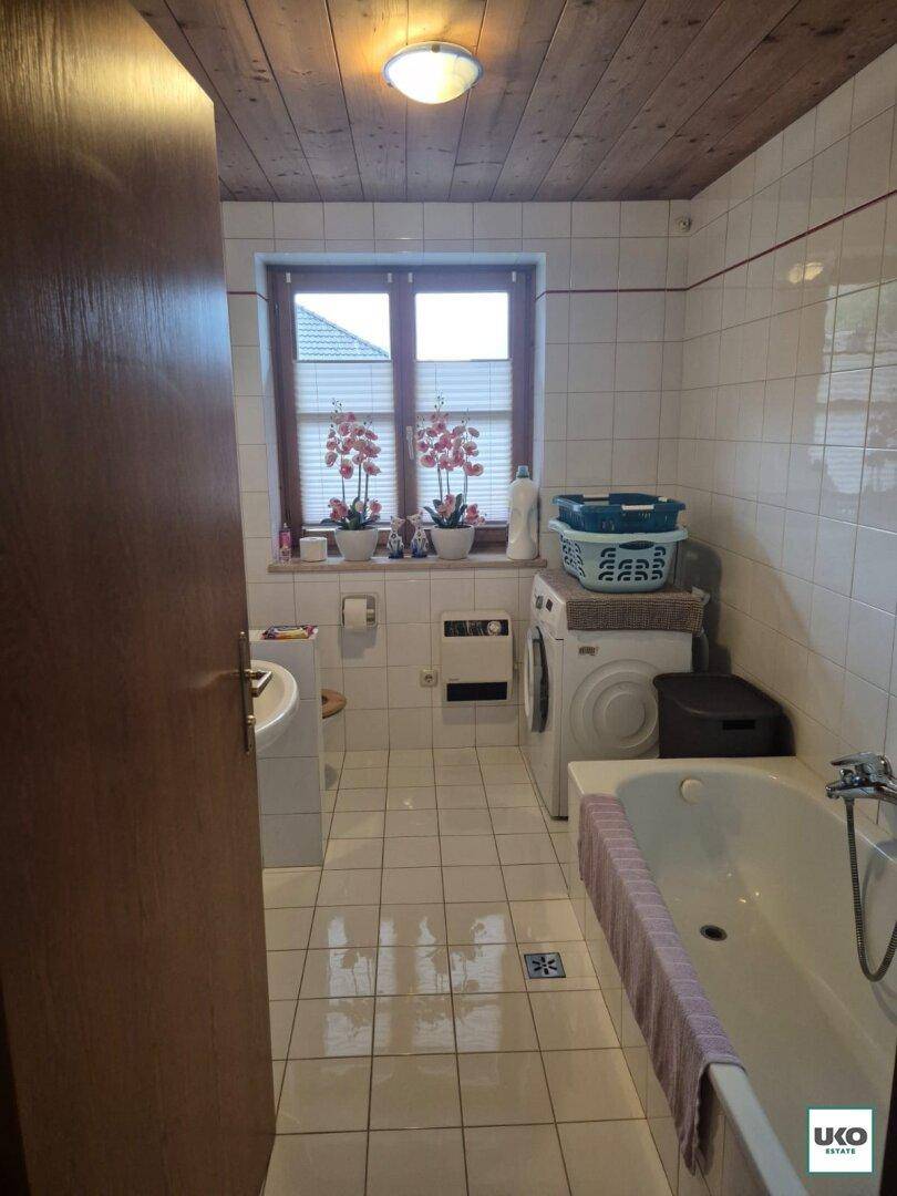 Helles Badezimmer mit weißen Fliesen, Badewanne, Fenster und integrierter Waschmaschine.