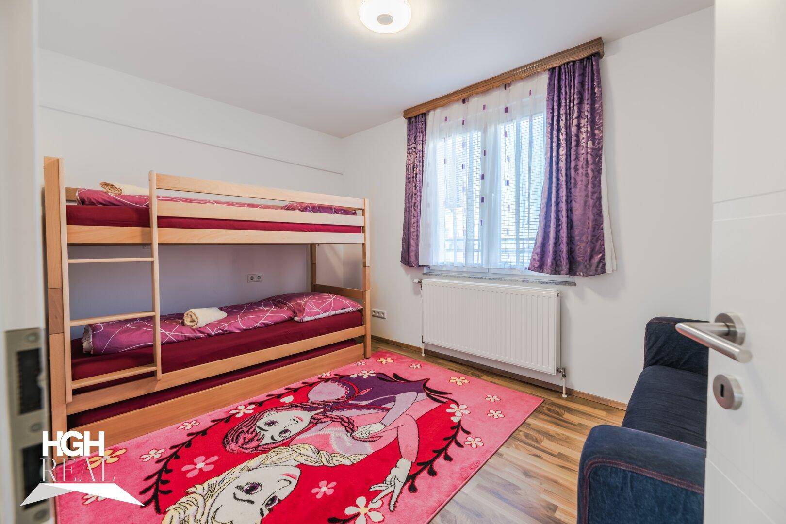 Helles Kinderzimmer mit Etagenbett, farbenfrohem Teppich und Blick nach draußen.