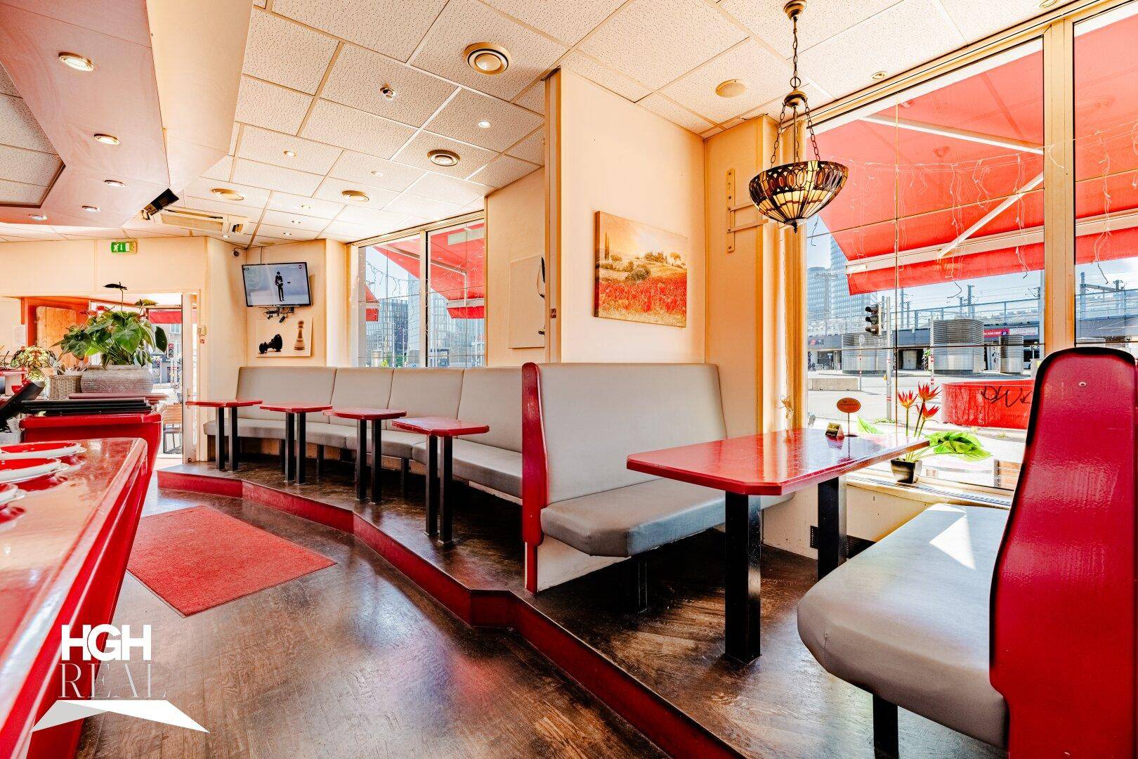 Innenansicht des Restaurants mit bequemen Sitzbänken, roten Tischen und großen Fenstern zur Straße.