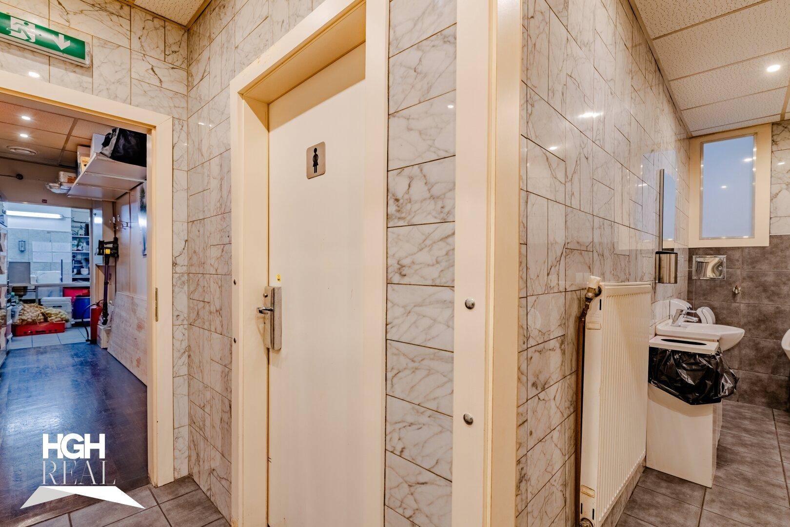 Eingangsbereich zu den Toiletten mit einer Tür, die das Damensymbol trägt, und Blick in einen Nebenraum.