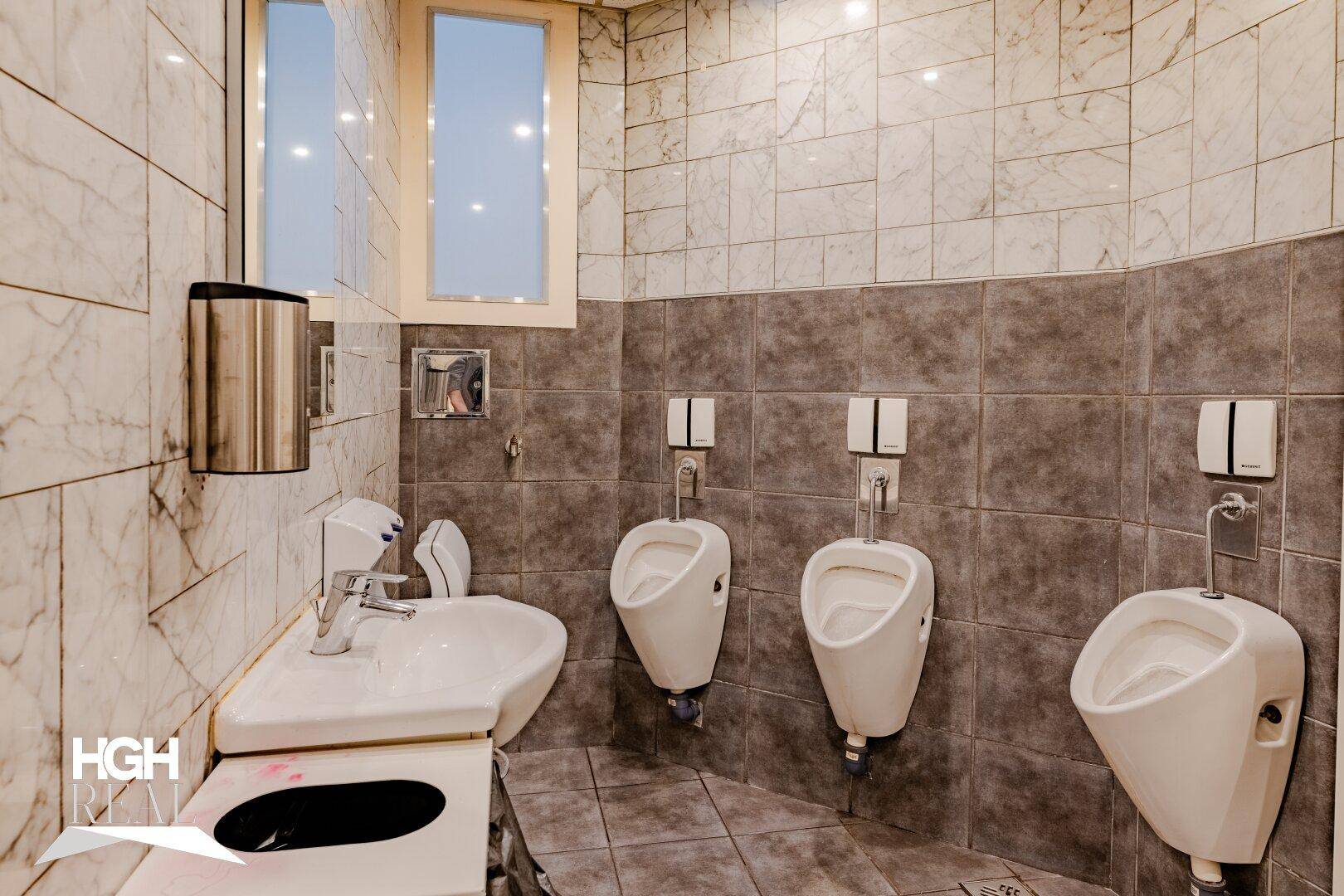 Herren-Waschraum mit drei Urinalen, einem Waschbecken und gefliesten Wänden in Grau und Marmoroptik.