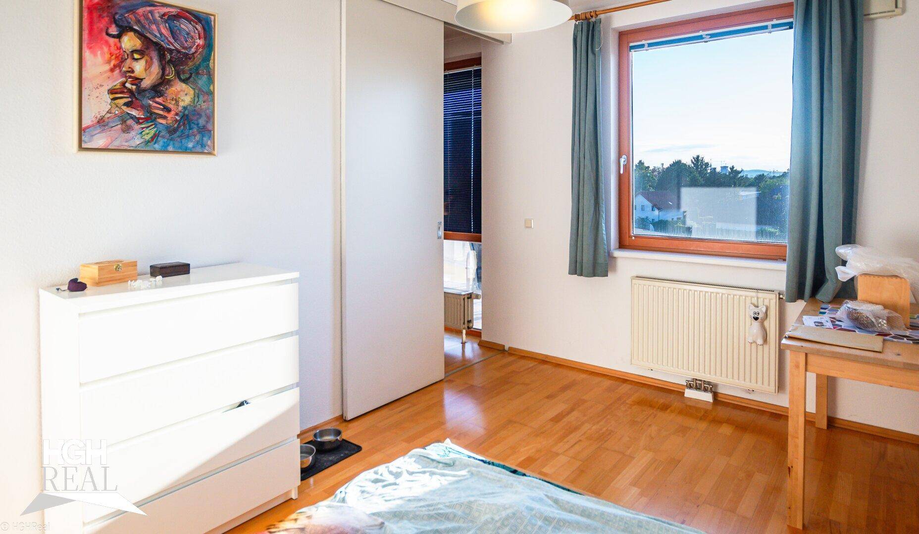 Schlafzimmer mit Holzboden, weißer Kommode und Fenster mit Ausblick ins Grüne.