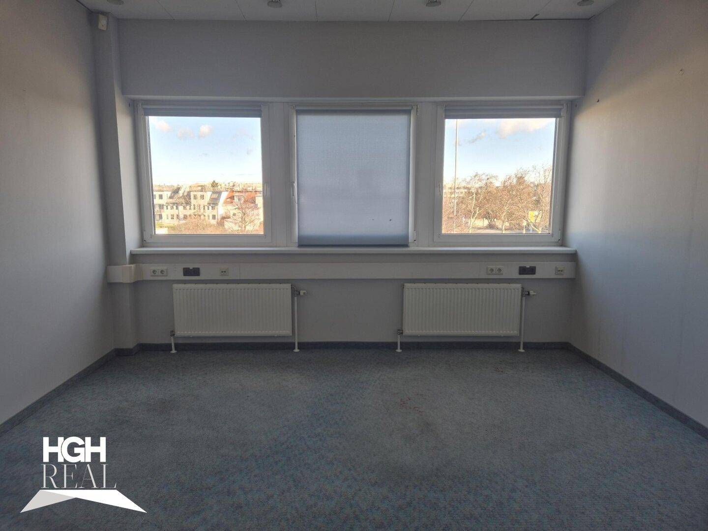 Leerer Büroraum mit Teppichboden und drei großen Fenstern, die eine weite Aussicht bieten.