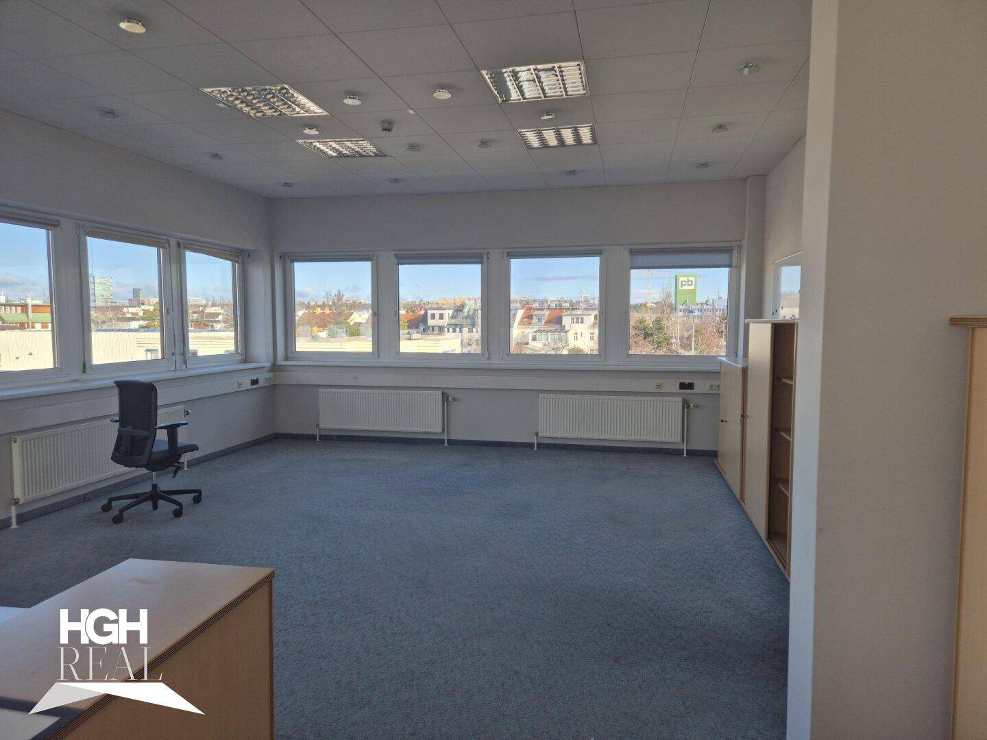 Großes Büro mit Teppichboden, vielen Fenstern und Blick auf die Stadt, mit einem Bürostuhl.