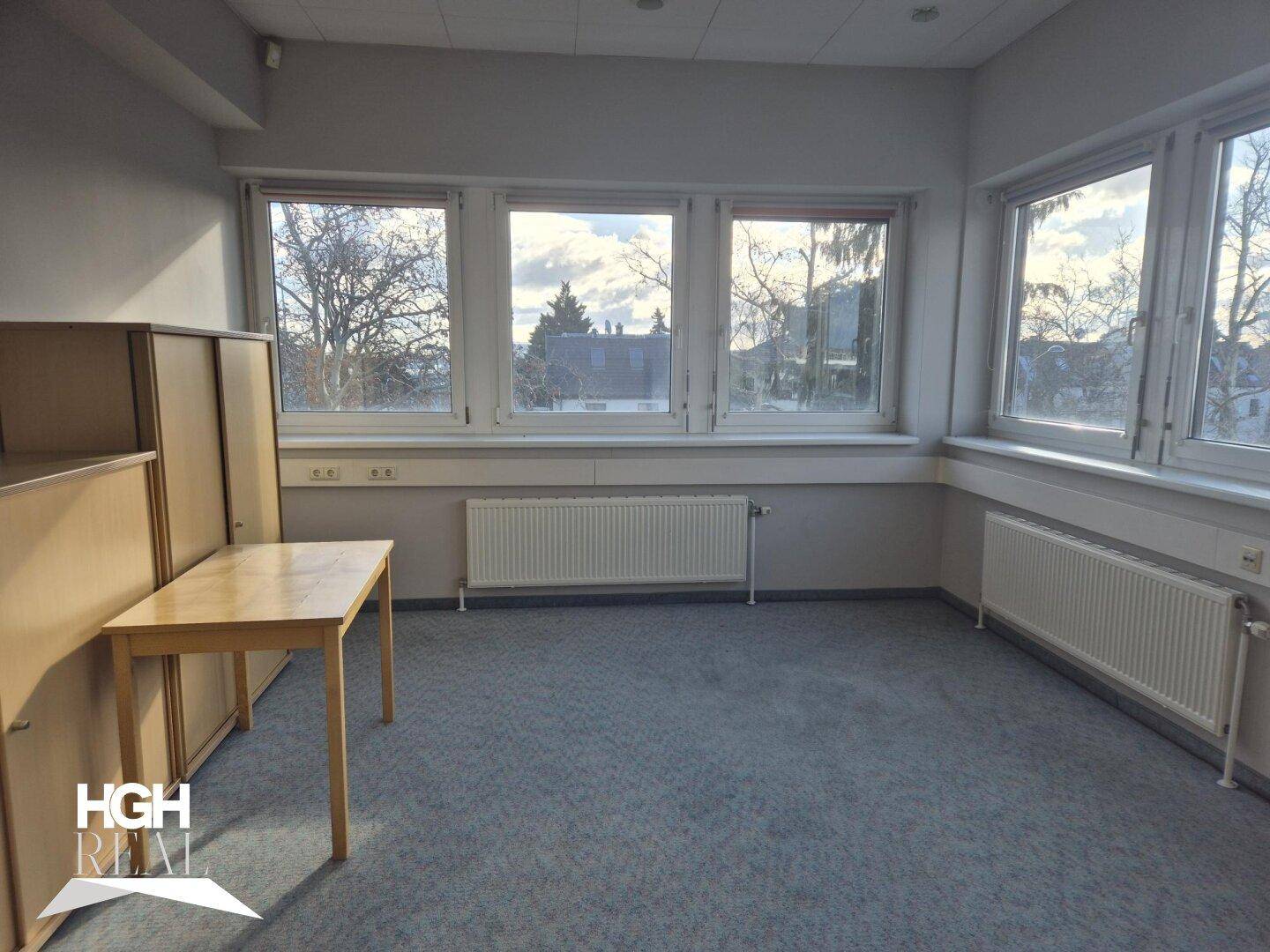 Helles Büro mit Teppichboden, großen Fenstern und Blick auf Bäume und Gebäude.