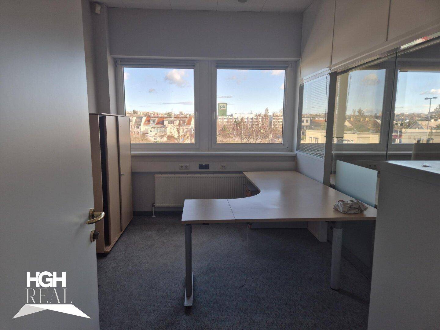Büro mit Teppichboden, Fenstern und einem L-förmigen Schreibtisch, bereit zur Nutzung.