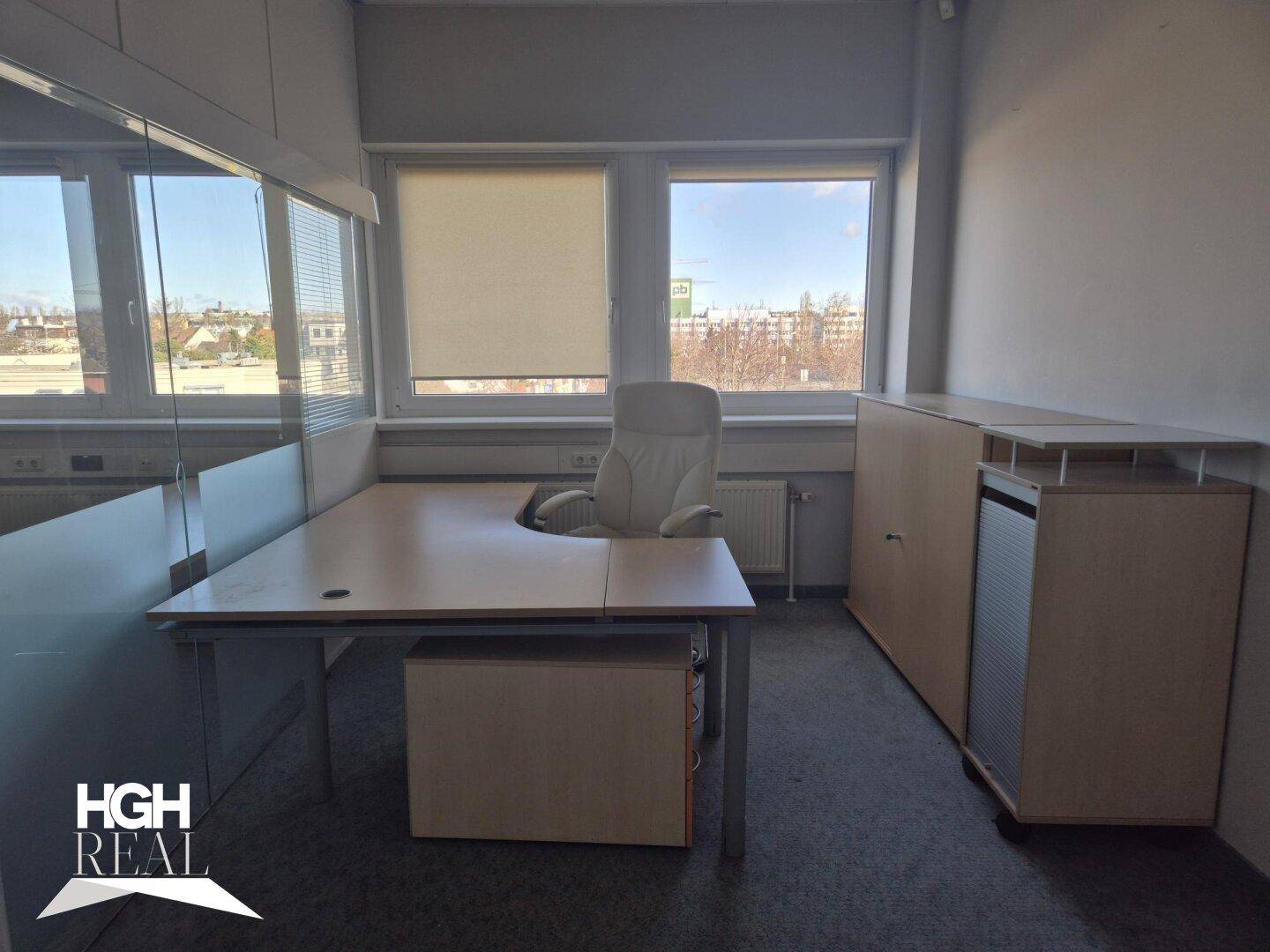 Büro mit Teppichboden, Fenstern und einem Schreibtisch mit Bürostuhl, gut beleuchtet.